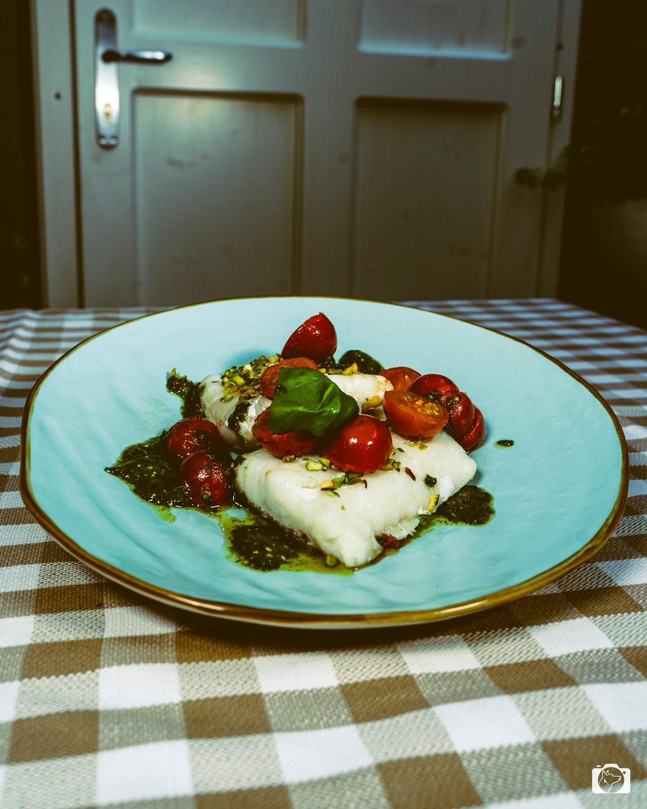 Piatto di pesce bianco, pomodorini e pesto su un piatto blu, adagiato su una tovaglia a quadri.