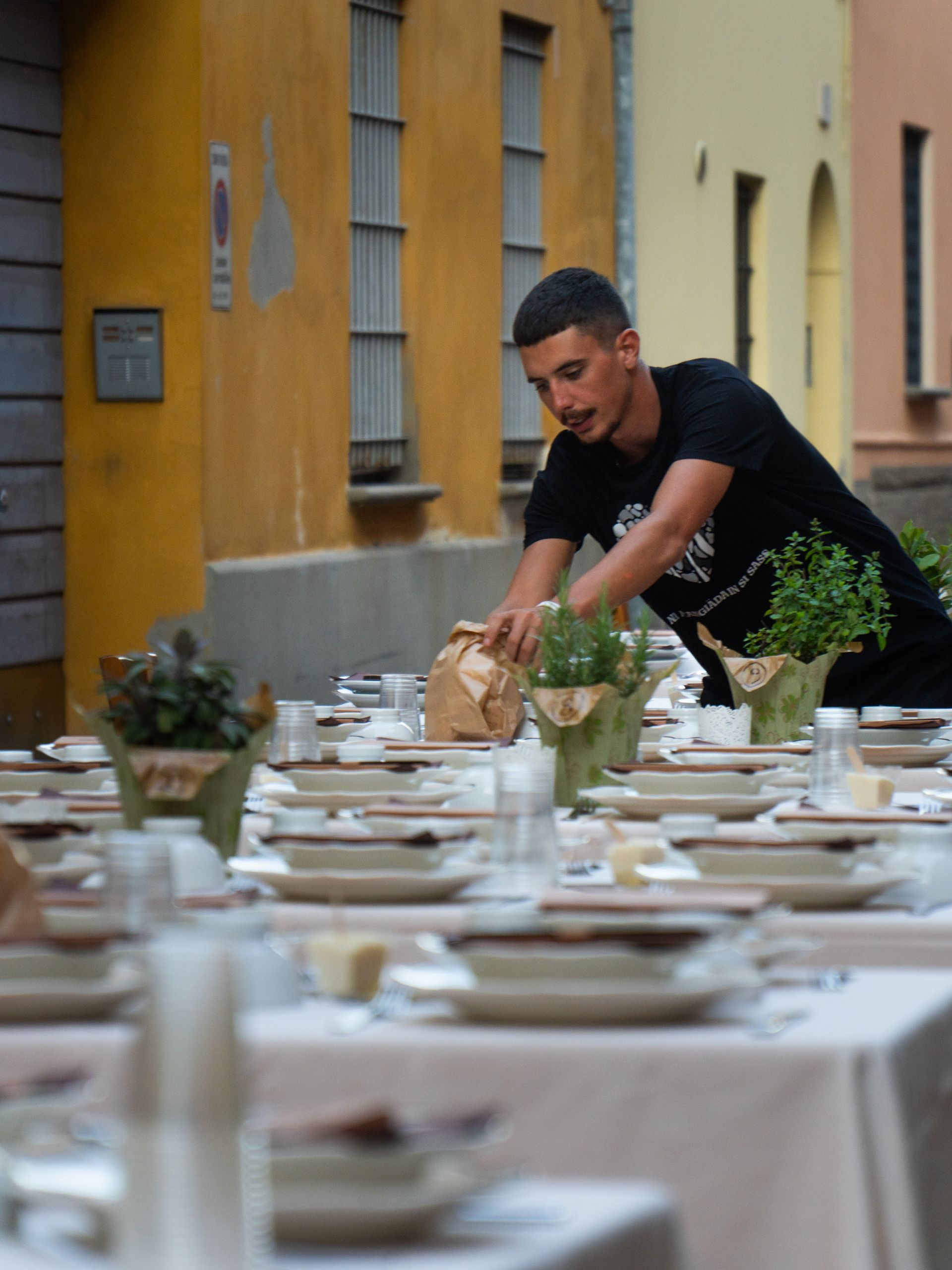 Uomo che apparecchia tavoli all'aperto con erbe aromatiche in vaso, vicino a edifici gialli e beige.