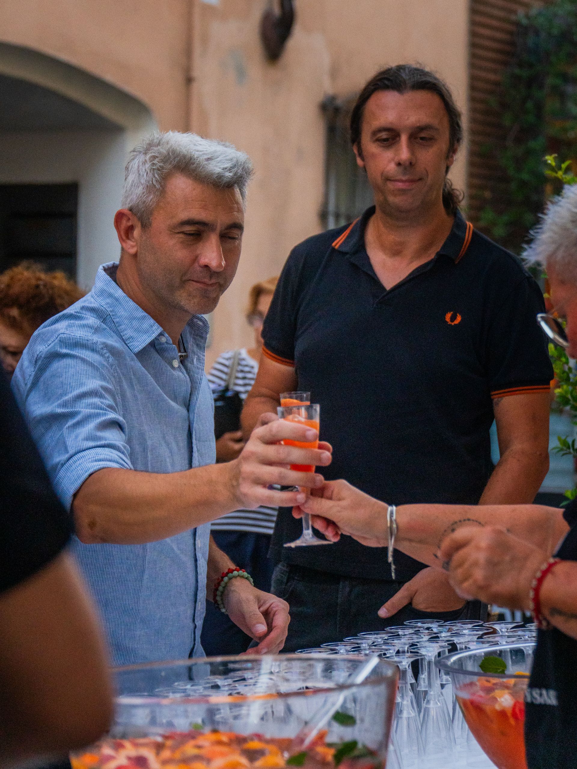 Un uomo con una camicia blu brinda con un cocktail, un altro uomo dietro di lui, evento all'aperto.
