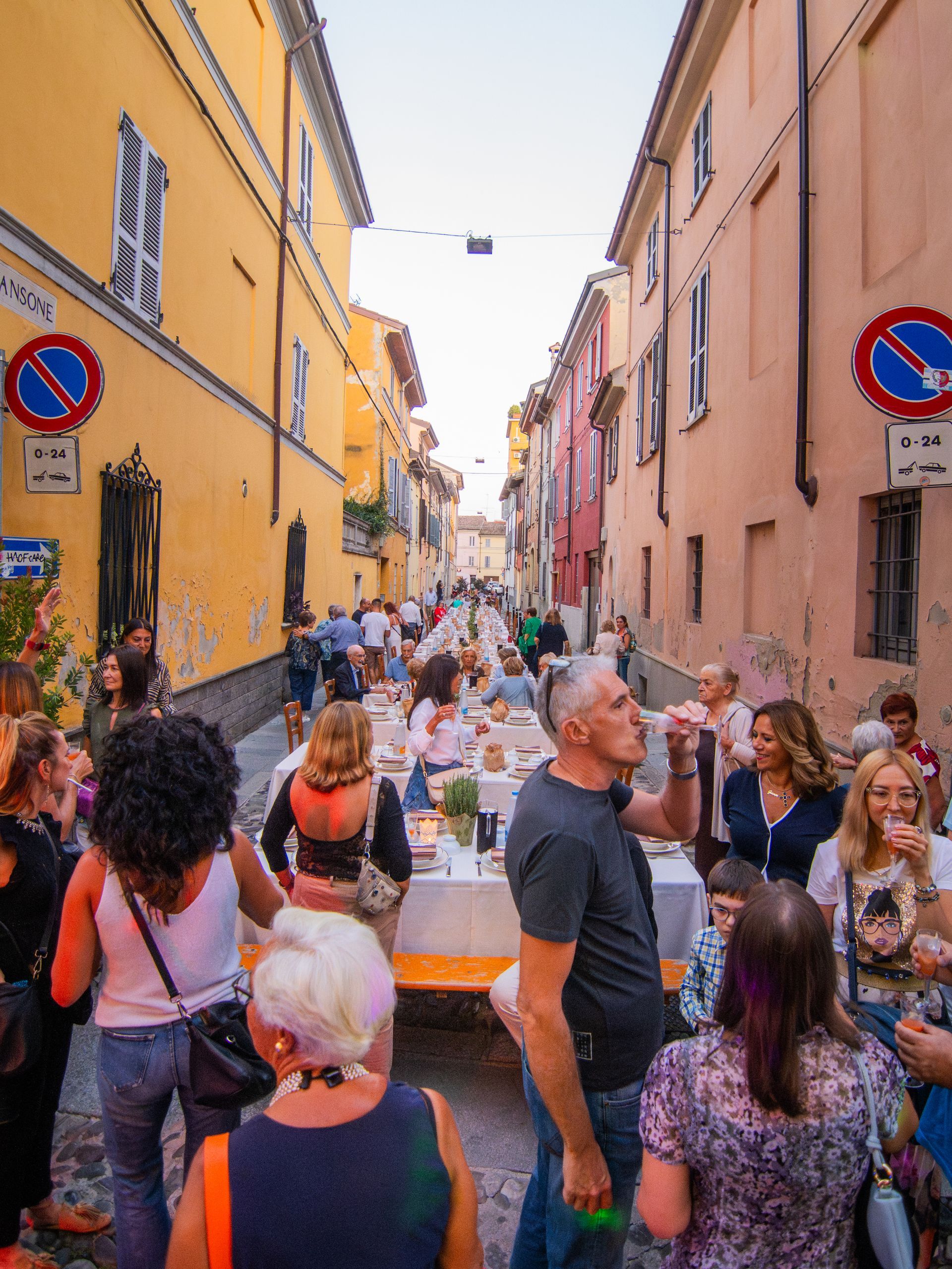 Un lungo tavolo apparecchiato per la cena in una stretta strada fiancheggiata da edifici. La gente si riunisce per mangiare e socializzare.