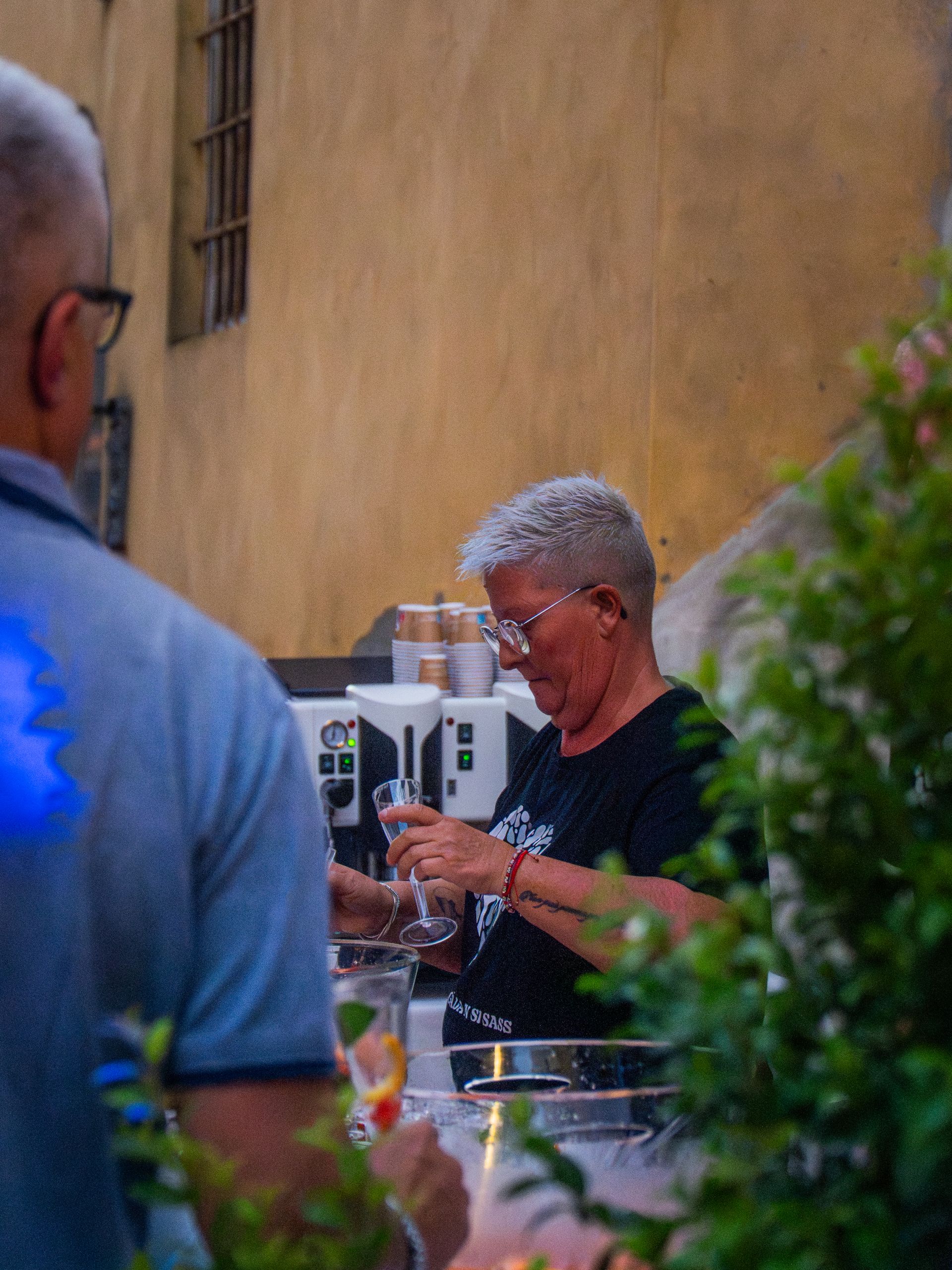 Una donna con i capelli grigi versa un drink dietro un bancone, un'altra persona in primo piano. Un edificio sullo sfondo.