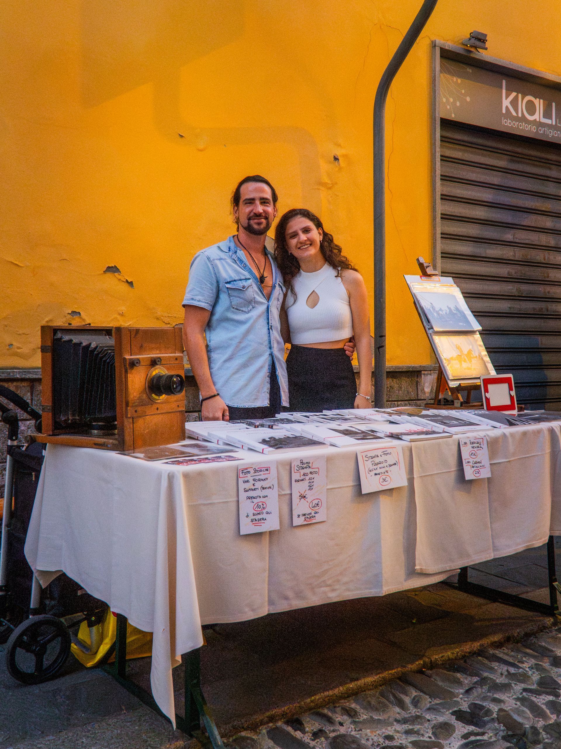 Due persone sorridenti davanti a un tavolo. Parete gialla sullo sfondo. Ambientazione stradale.
