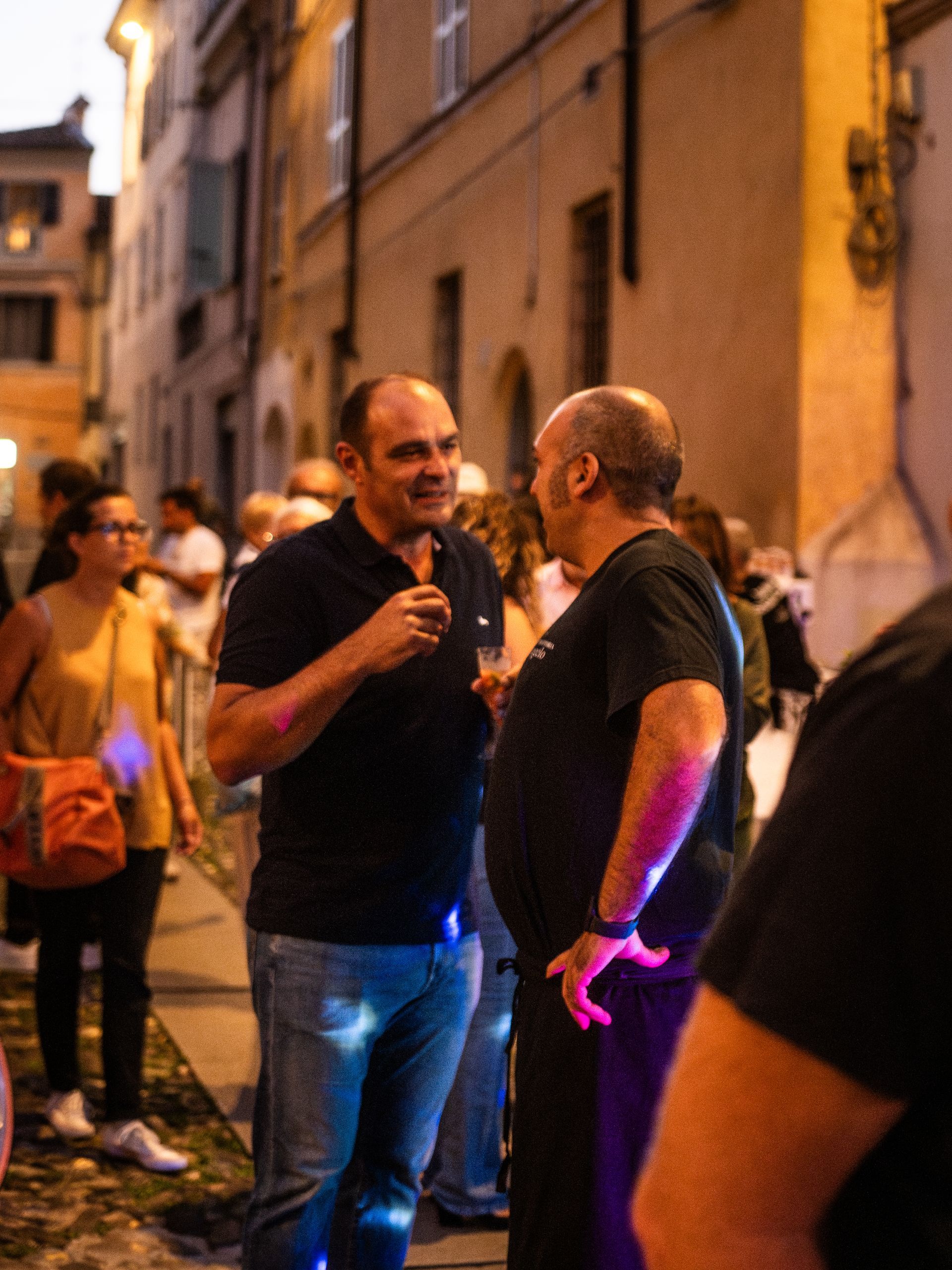 Due uomini conversano in una strada cittadina di notte; uno ha un drink in mano. Sullo sfondo si vedono altre persone ed edifici.
