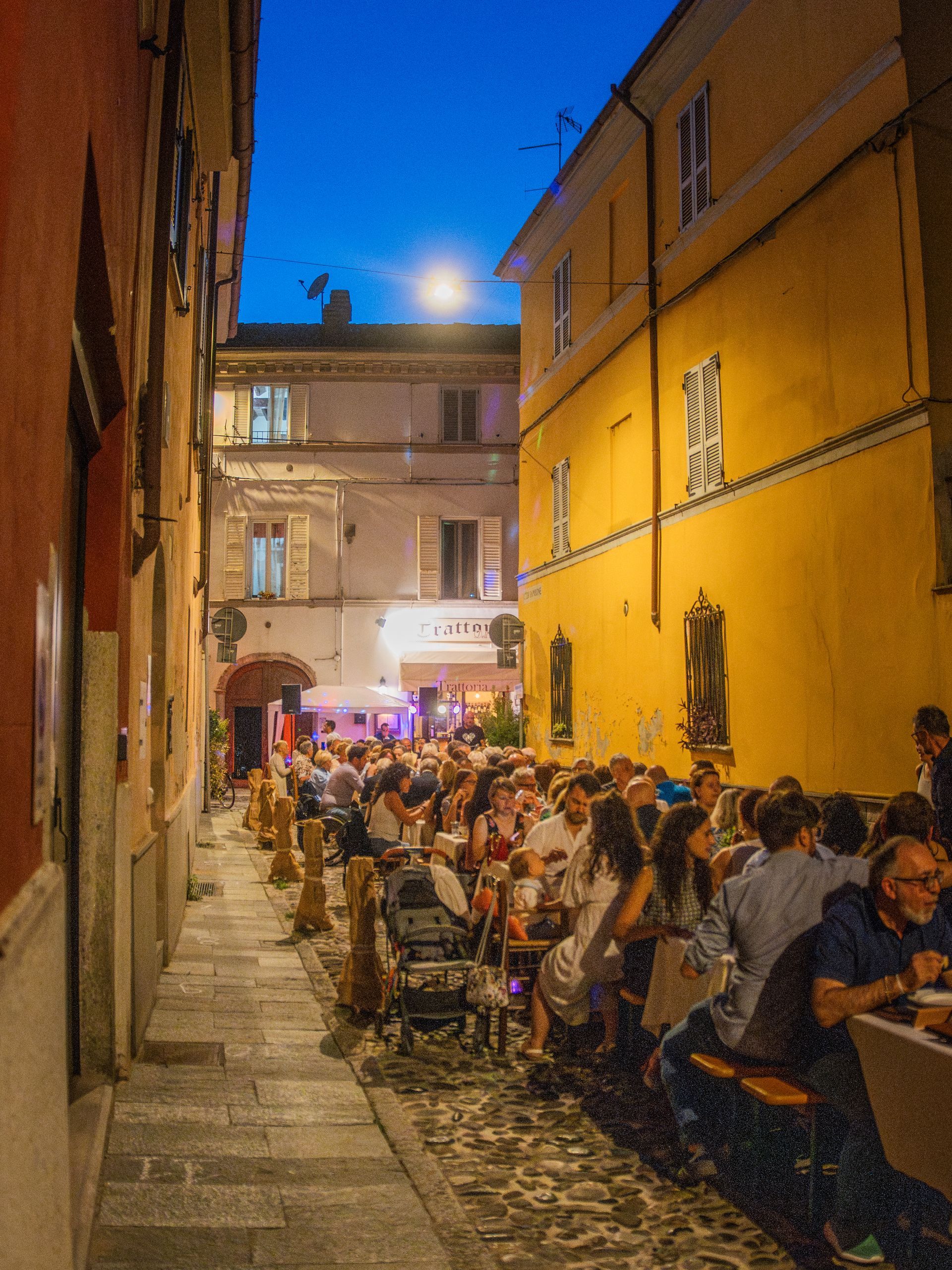 Stretta strada acciottolata con ristorante all'aperto affollato al tramonto, illuminato dai lampioni.