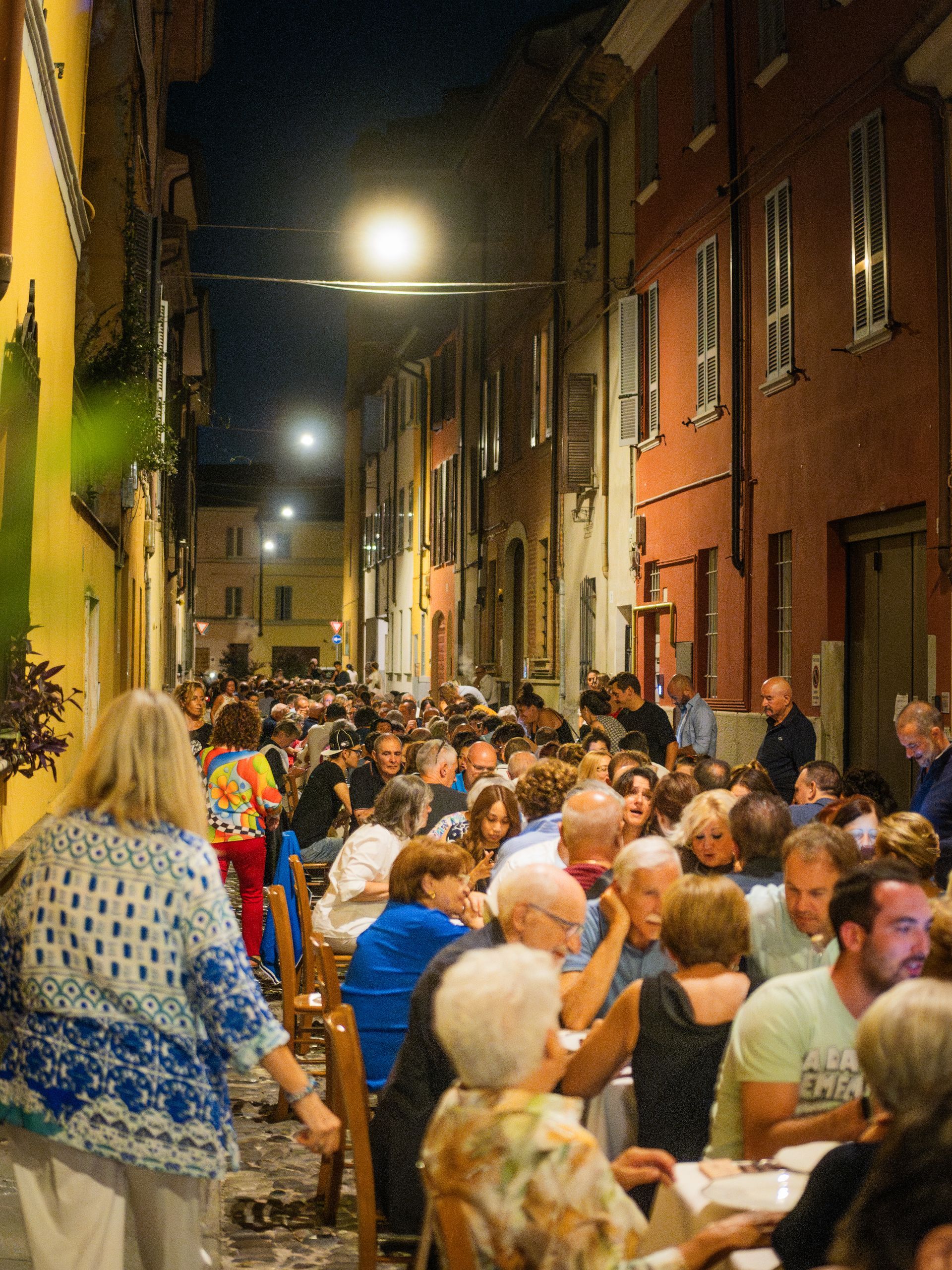 Persone che cenano all'aperto di notte, in una stretta strada europea fiancheggiata da edifici e lampioni.