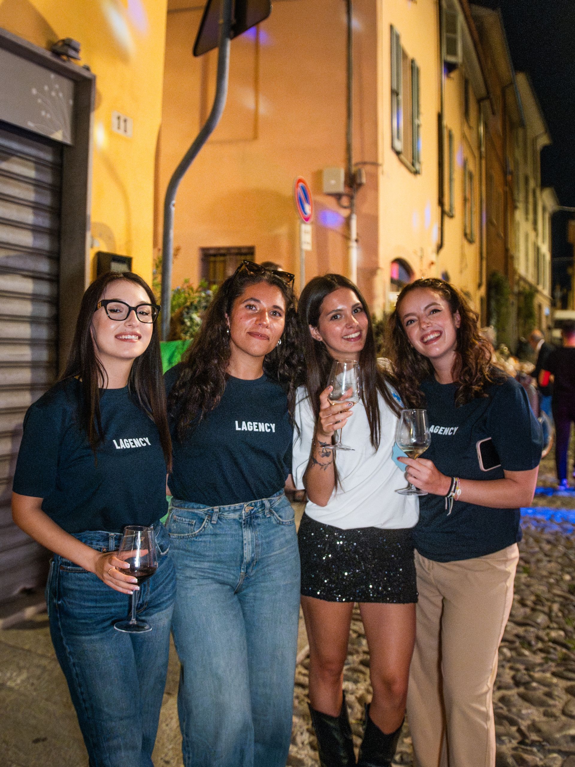 Quattro persone, sorridenti, con bicchieri di vino, posano all'aperto di notte. Si vedono edifici e ciottoli.