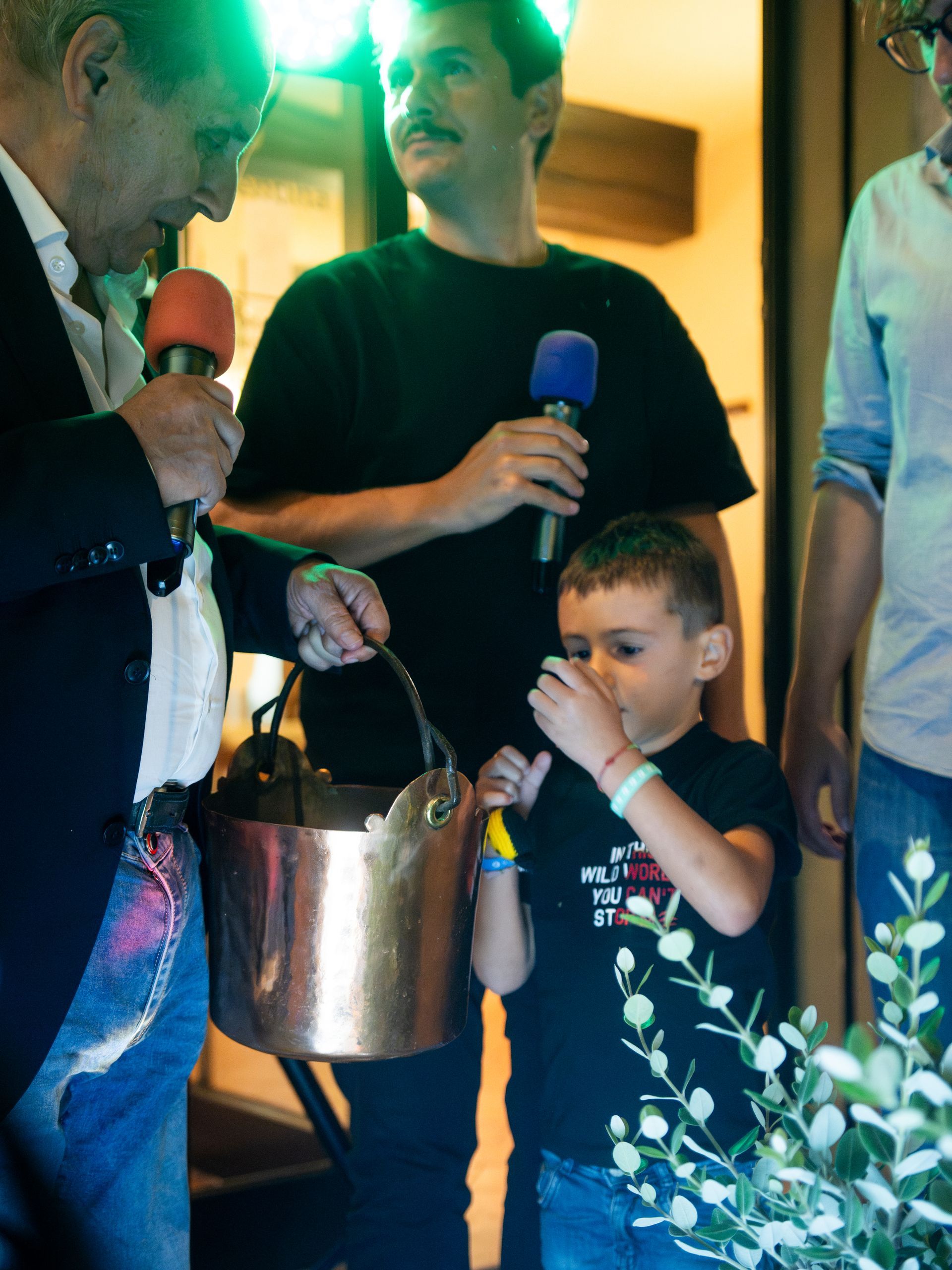 Persone che tengono microfoni vicino a un secchio di rame, probabilmente durante un evento, con un bambino piccolo.