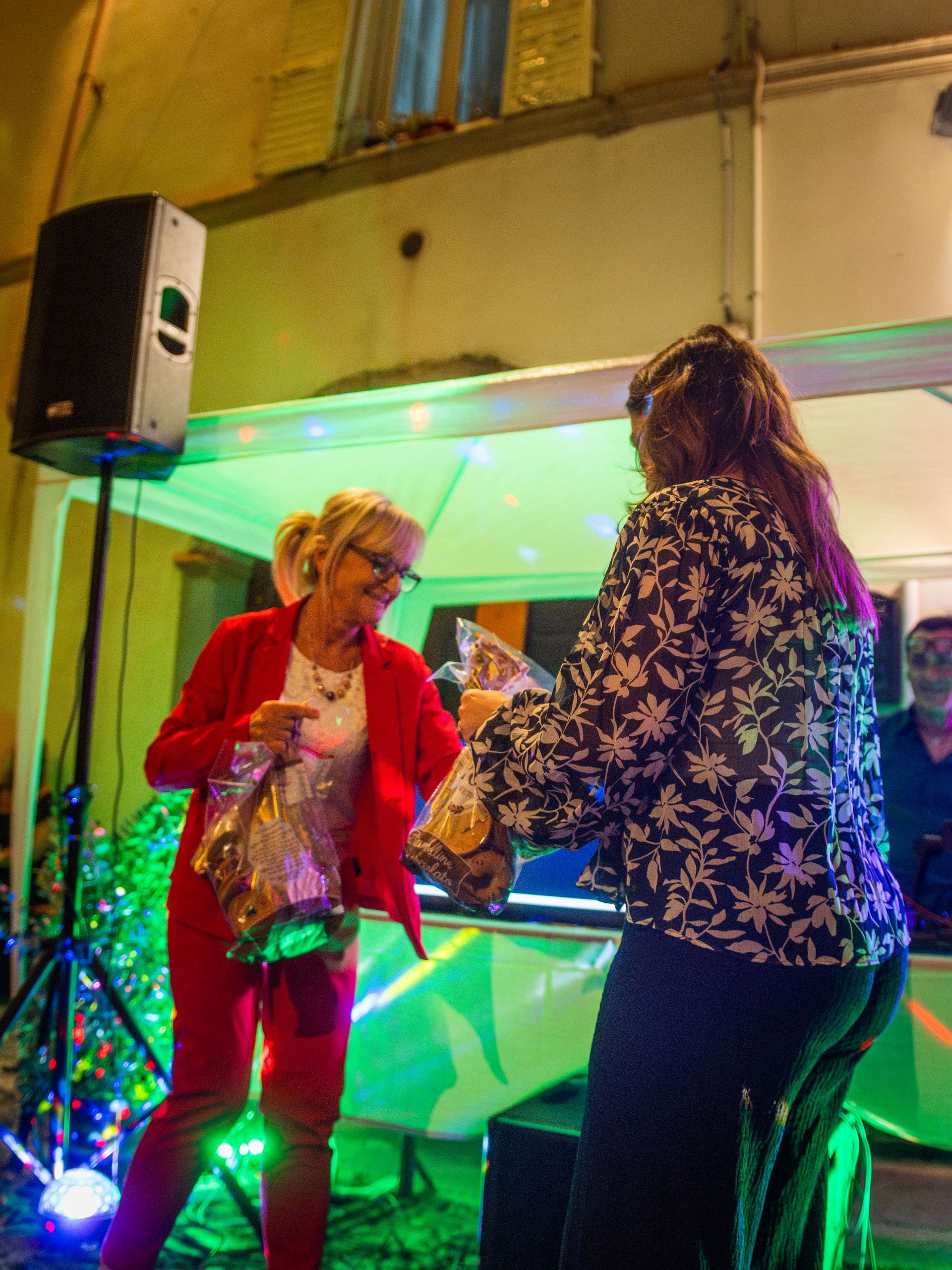 Due persone si scambiano regali vicino a una cabina da DJ decorata all'aperto, di notte. Una indossa un abito rosso, l'altra una giacca a fiori.