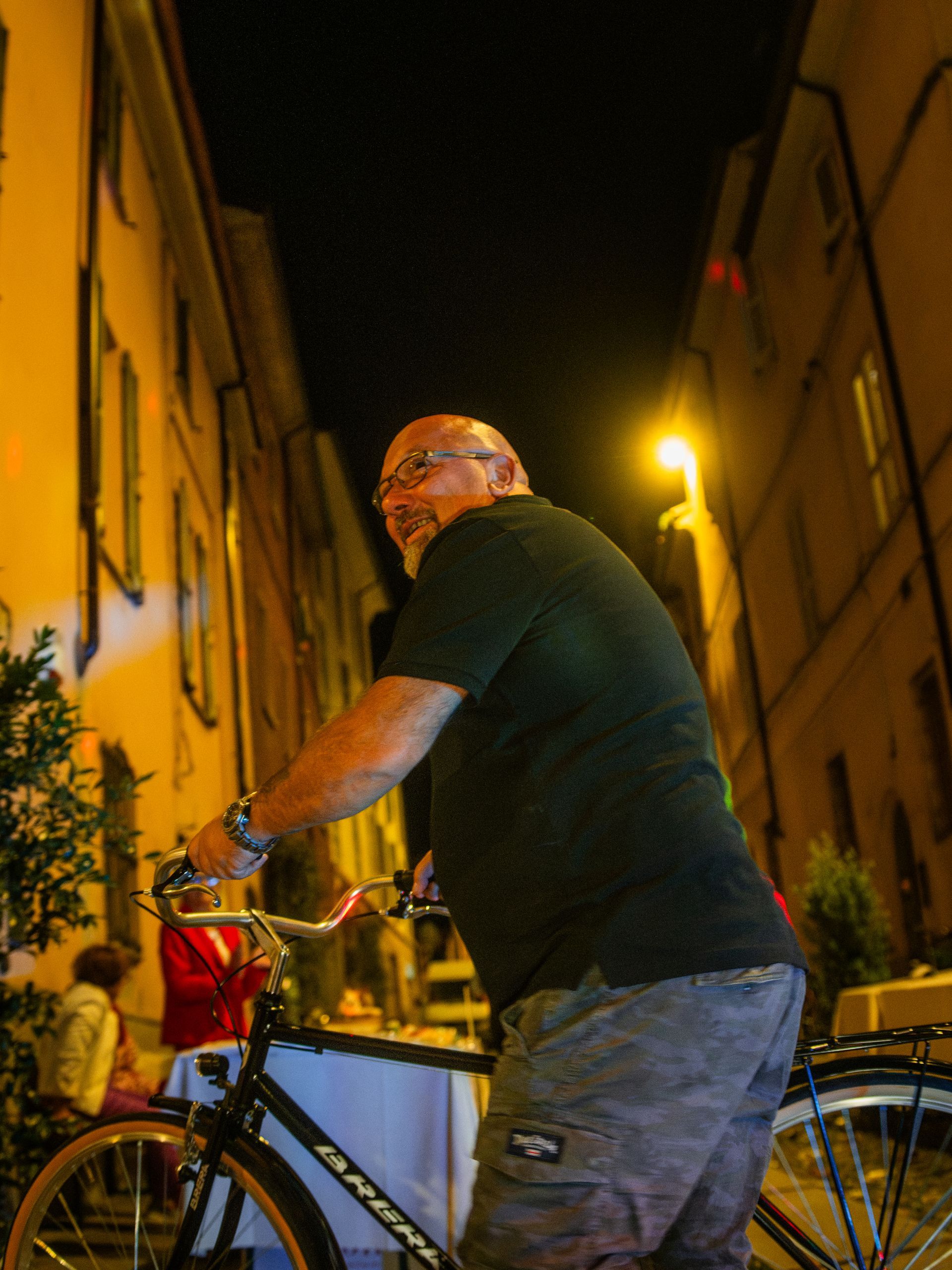 Uomo in bicicletta in una strada stretta di notte, sorridente. Gli edifici si allineano su entrambi i lati.