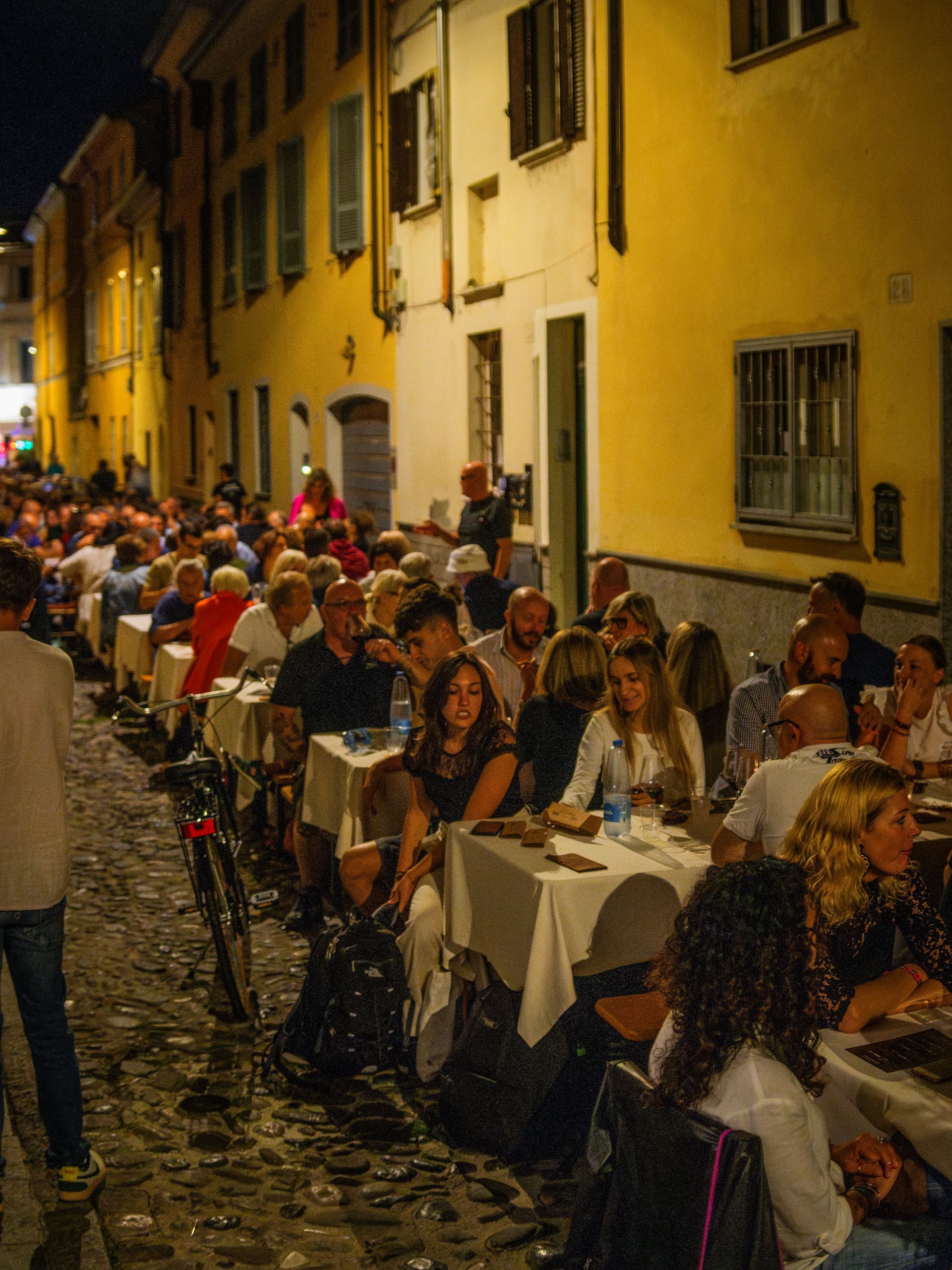 Ristorante all'aperto di notte: persone sedute a lunghi tavoli, illuminate dai lampioni, in una stretta strada acciottolata.