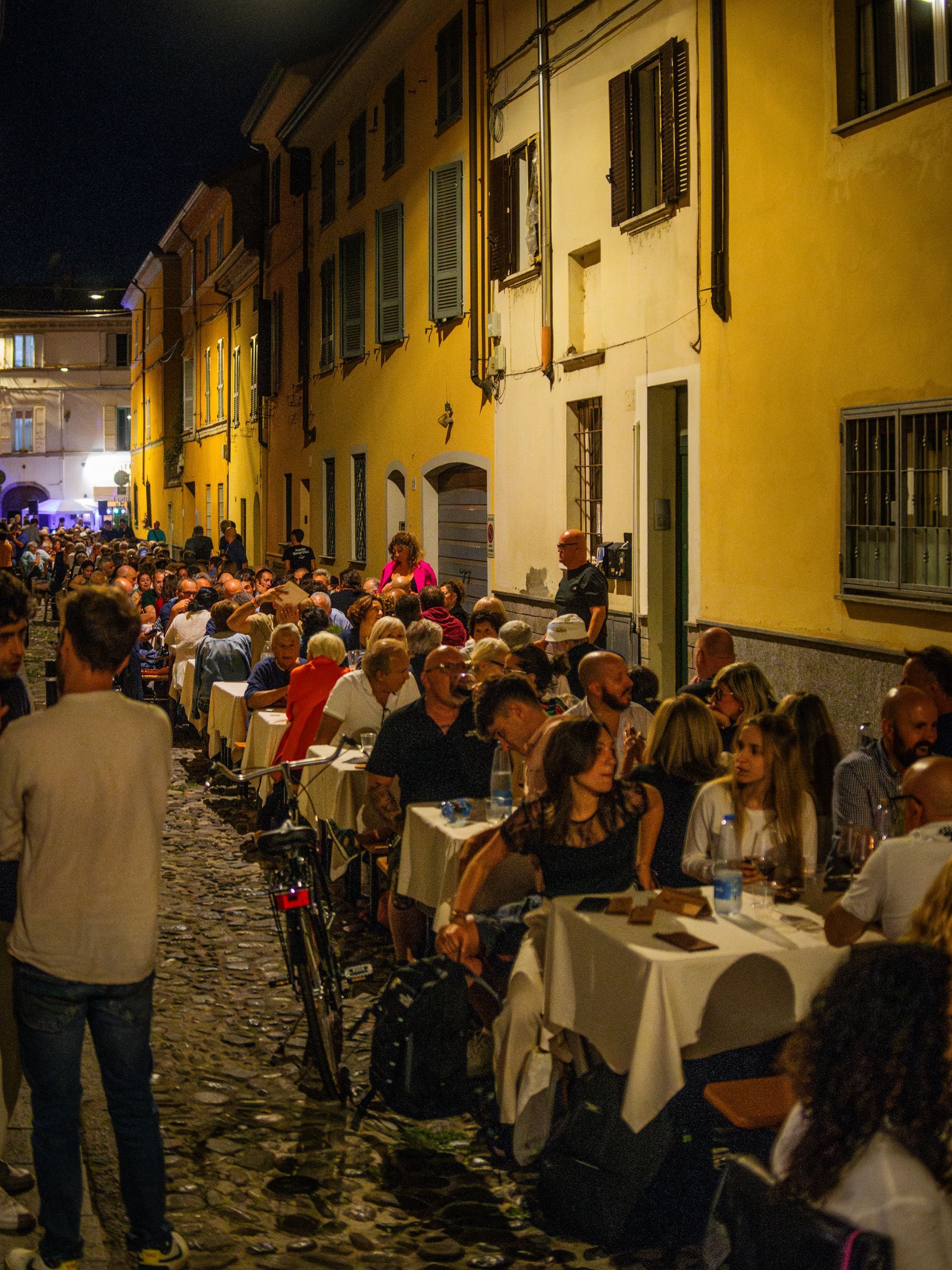 Scena di strada notturna; cena all'aperto con molte persone sedute ai tavoli lungo gli edifici gialli.