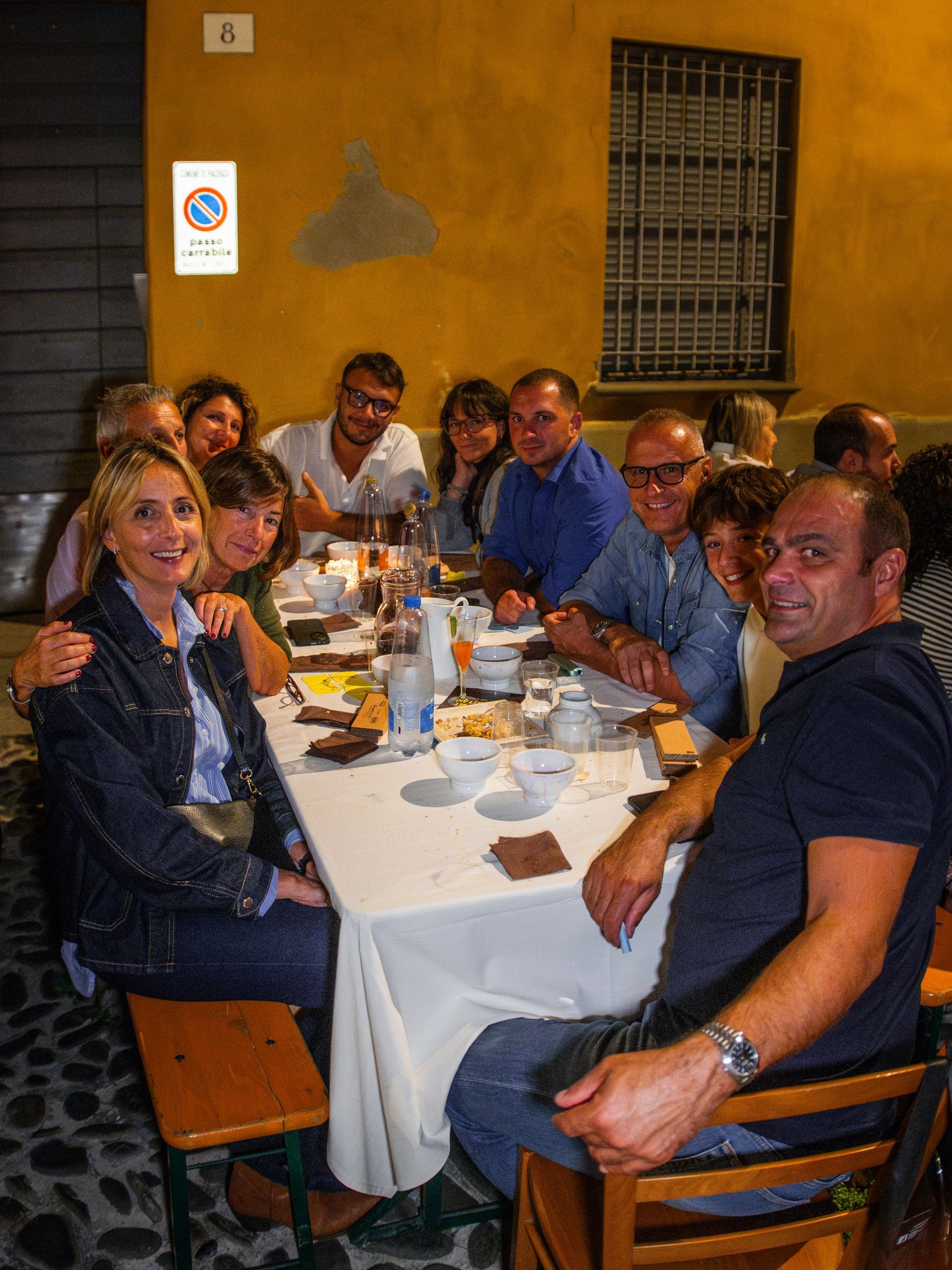 Gruppo di persone sedute attorno a un tavolo, all'aperto, sorridenti. Muro giallo, cartello stradale.