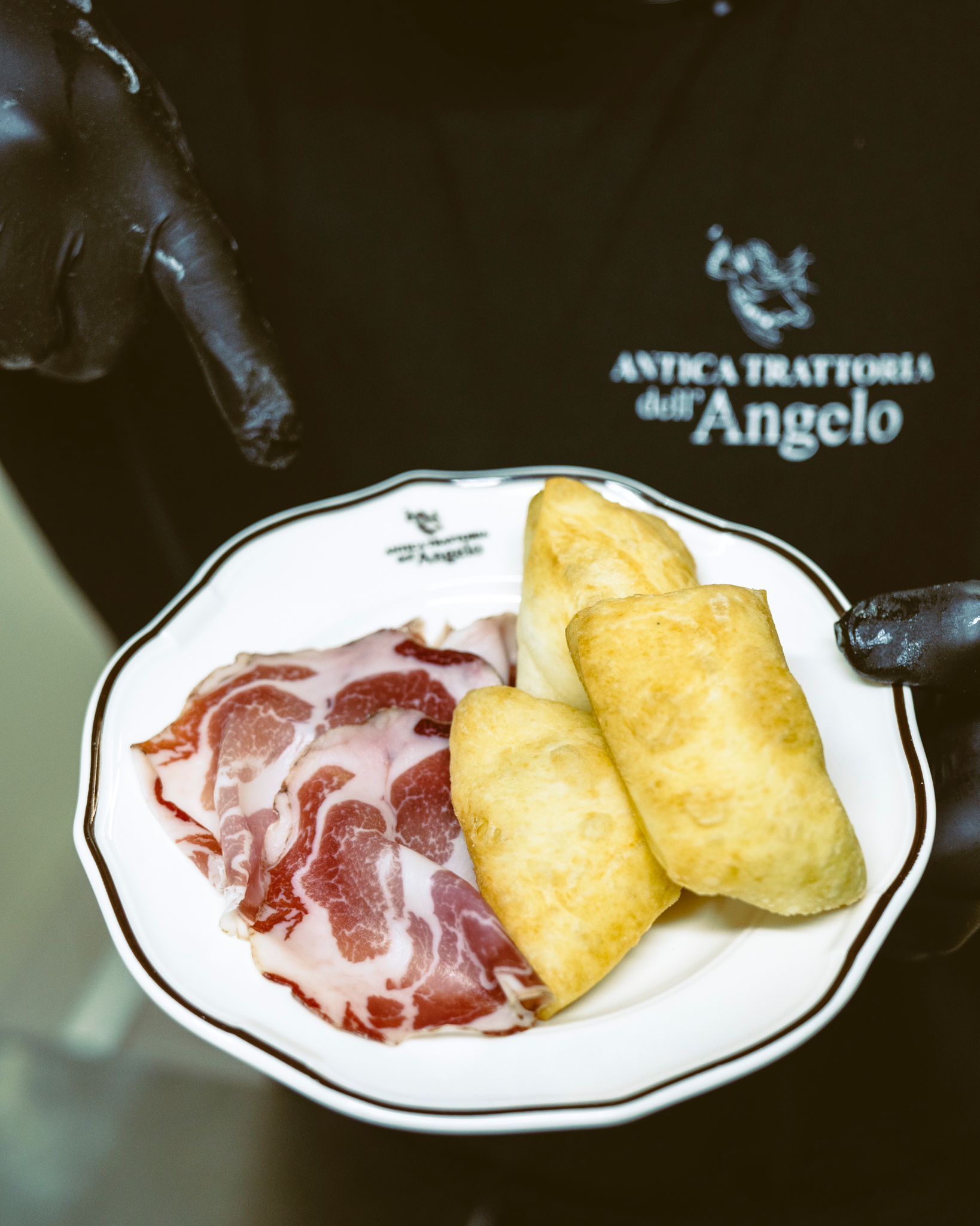 Piatto di carne affettata e pasta fritta tenuto da una persona con guanti neri, logo del ristorante visibile.
