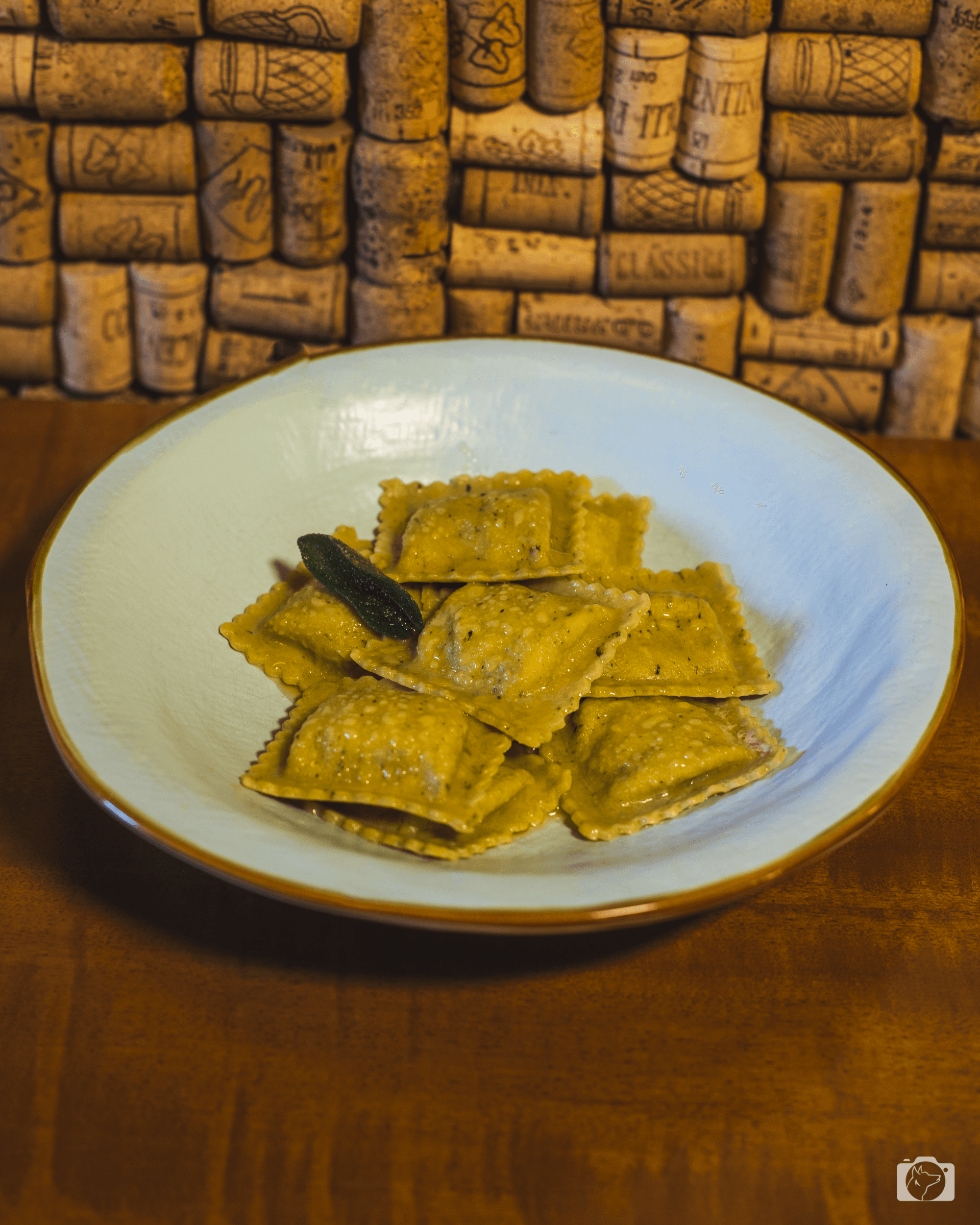 Ravioli su un piatto bianco con bordo marrone, guarnito con una foglia di salvia fritta, davanti a uno sfondo di sughero.