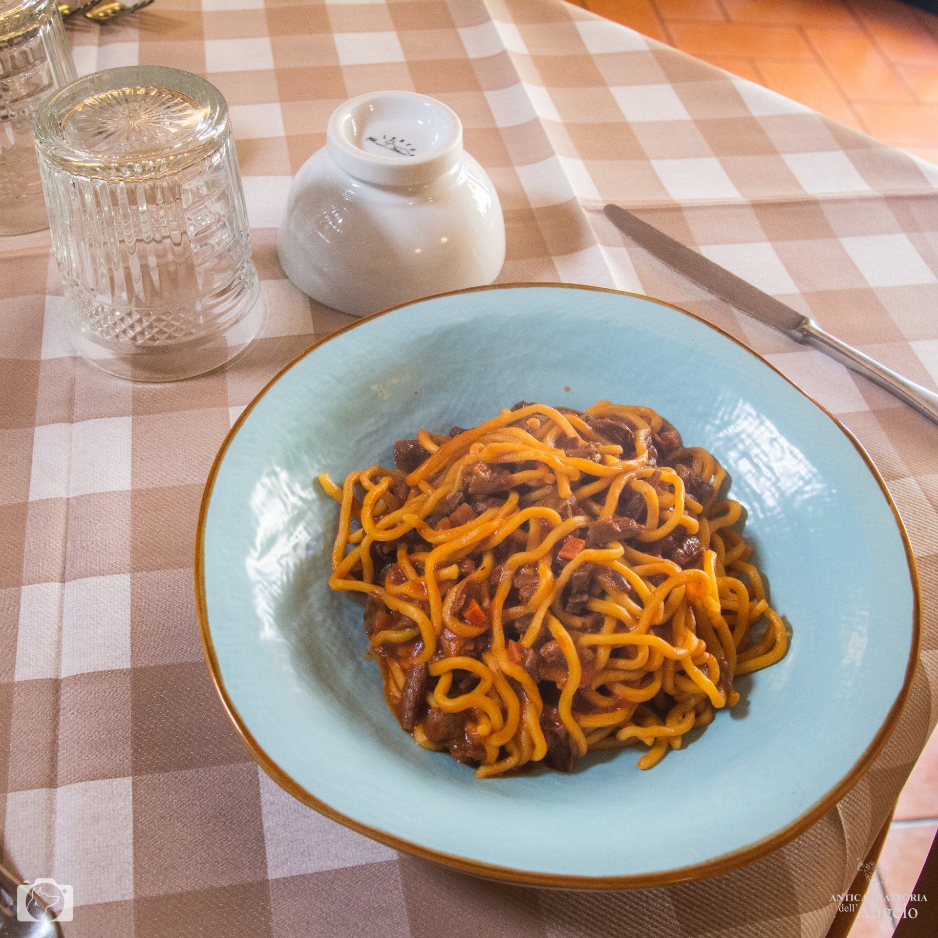 Spaghetti al ragù su un piatto blu, adagiati su una tovaglia a quadri, con un bicchiere, una saliera e un coltello.