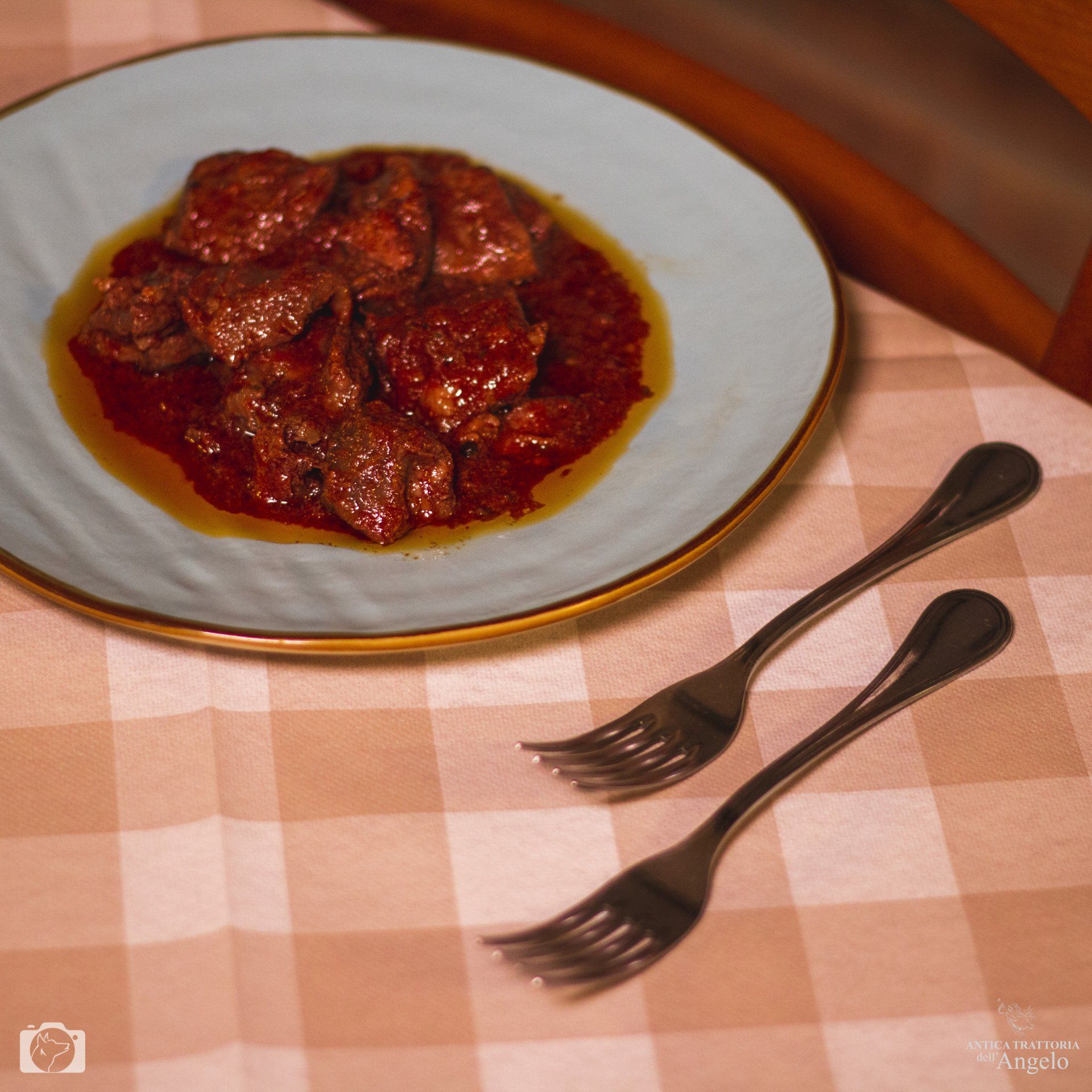 Carne stufata in salsa rossa su un piatto blu, due forchette su una tovaglia a quadri.