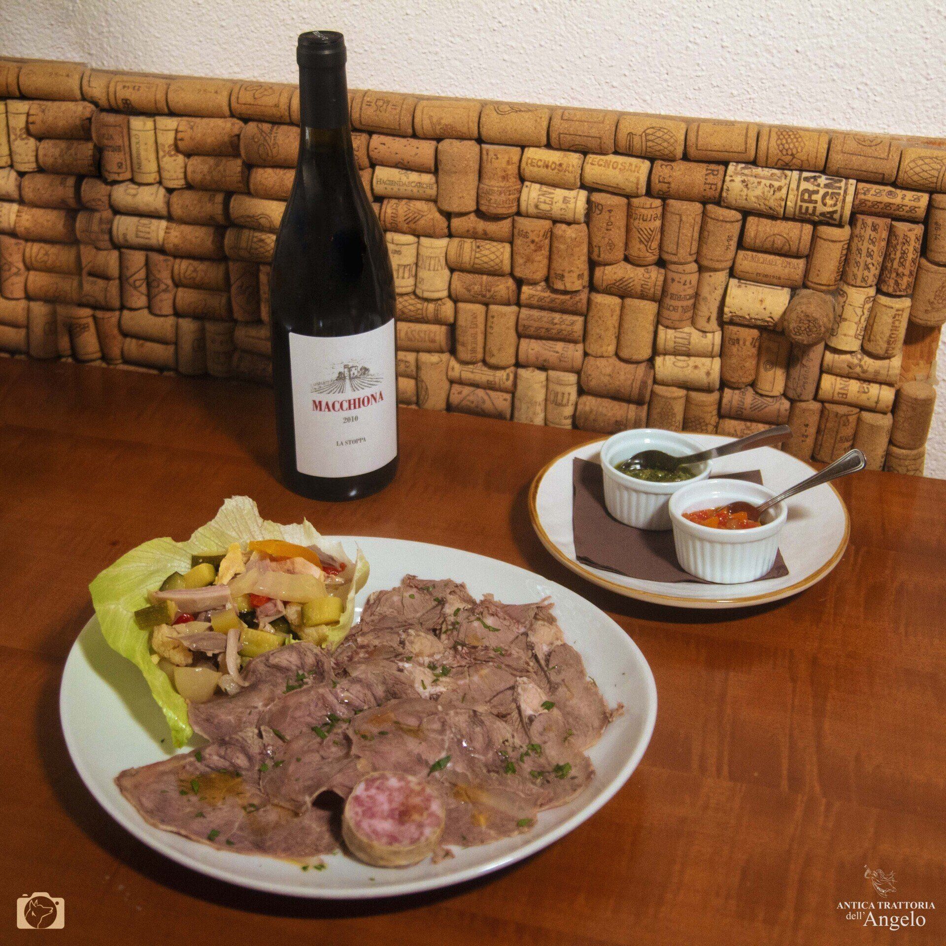 Piatto di carne affettata con contorno di insalata, salse e bottiglia di vino su un tavolo con una parete di sughero.