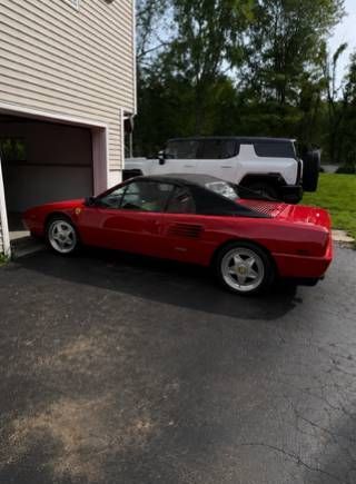 Red sports car parked beside a house driveway, with a white SUV behind it.