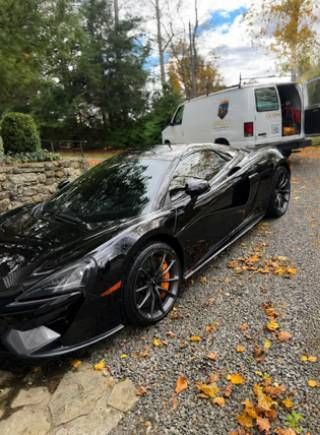 Black sports car parked on a leaf-covered road beside a white van and stone wall