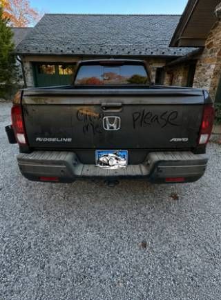 Black Honda Ridgeline parked in a driveway in front of a house, with a handwritten “Please” on the tailgate.