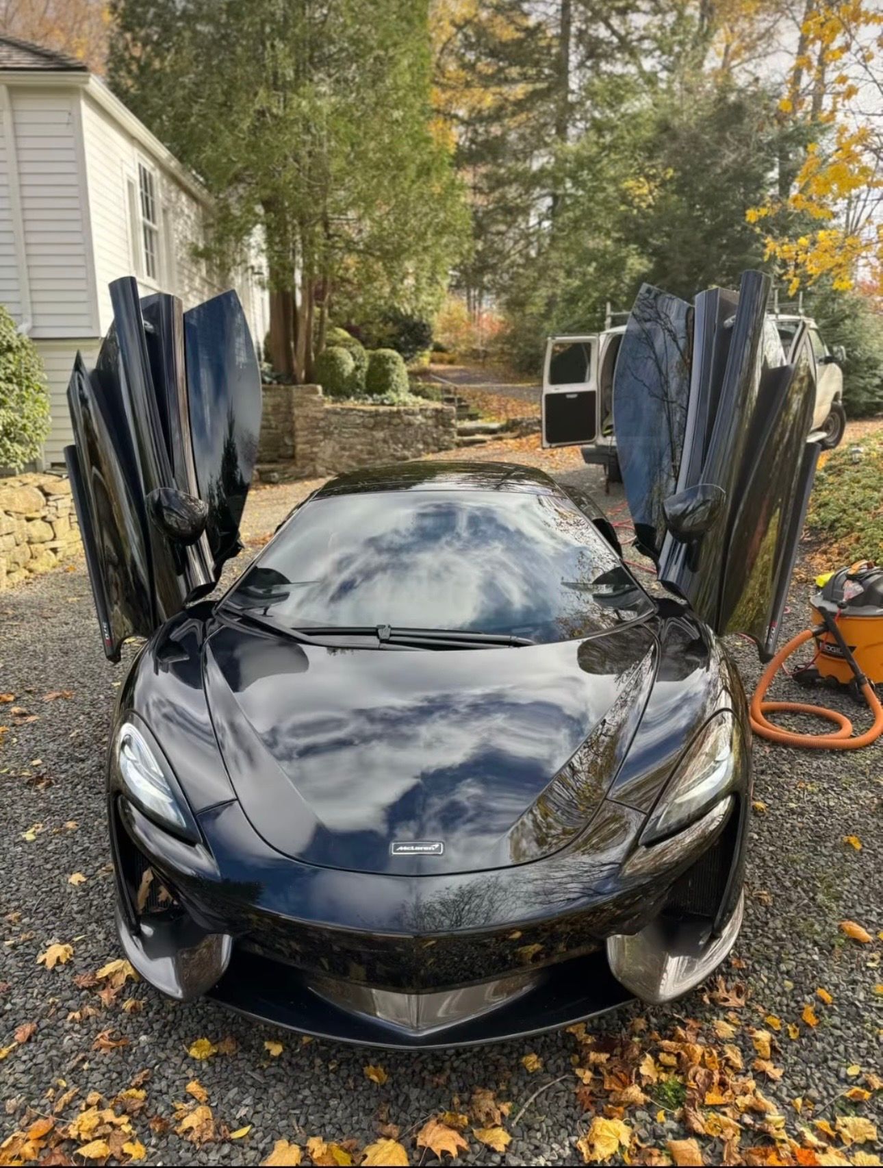 Black sports car with butterfly doors open, parked in a driveway with autumn leaves.