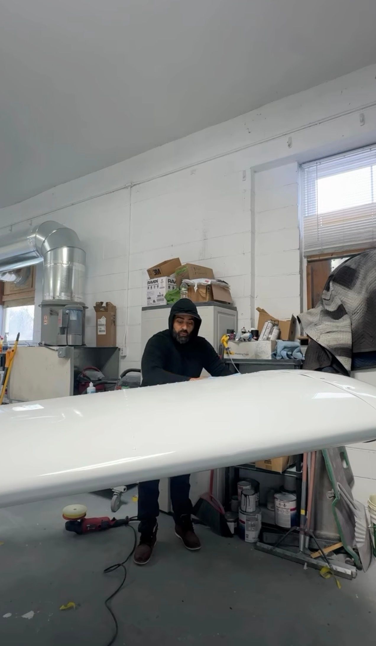 Person in a workshop stands behind a long white object amid shelves and tools.