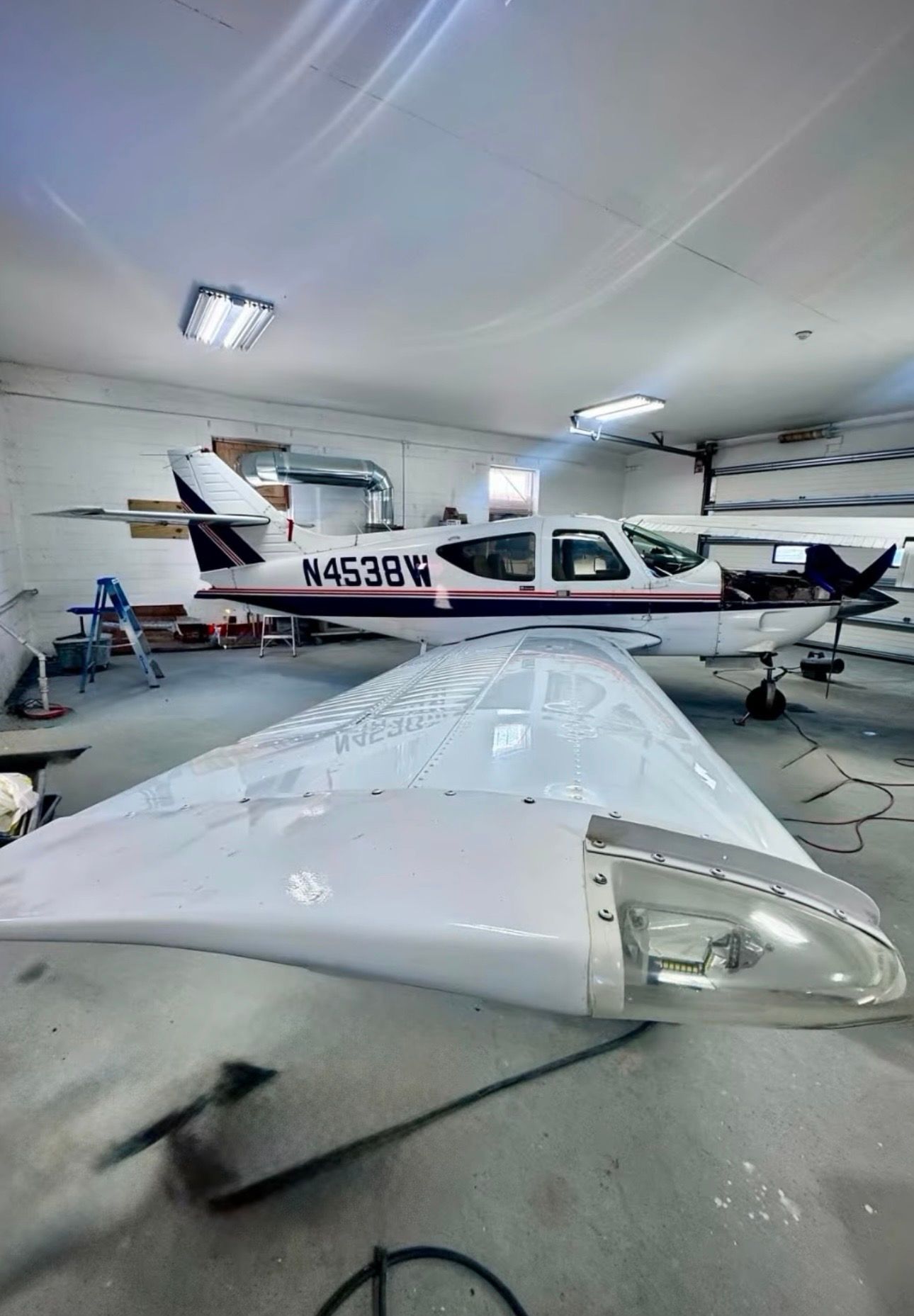 Small white airplane in a hangar, viewed from the wing with another plane in the background.
