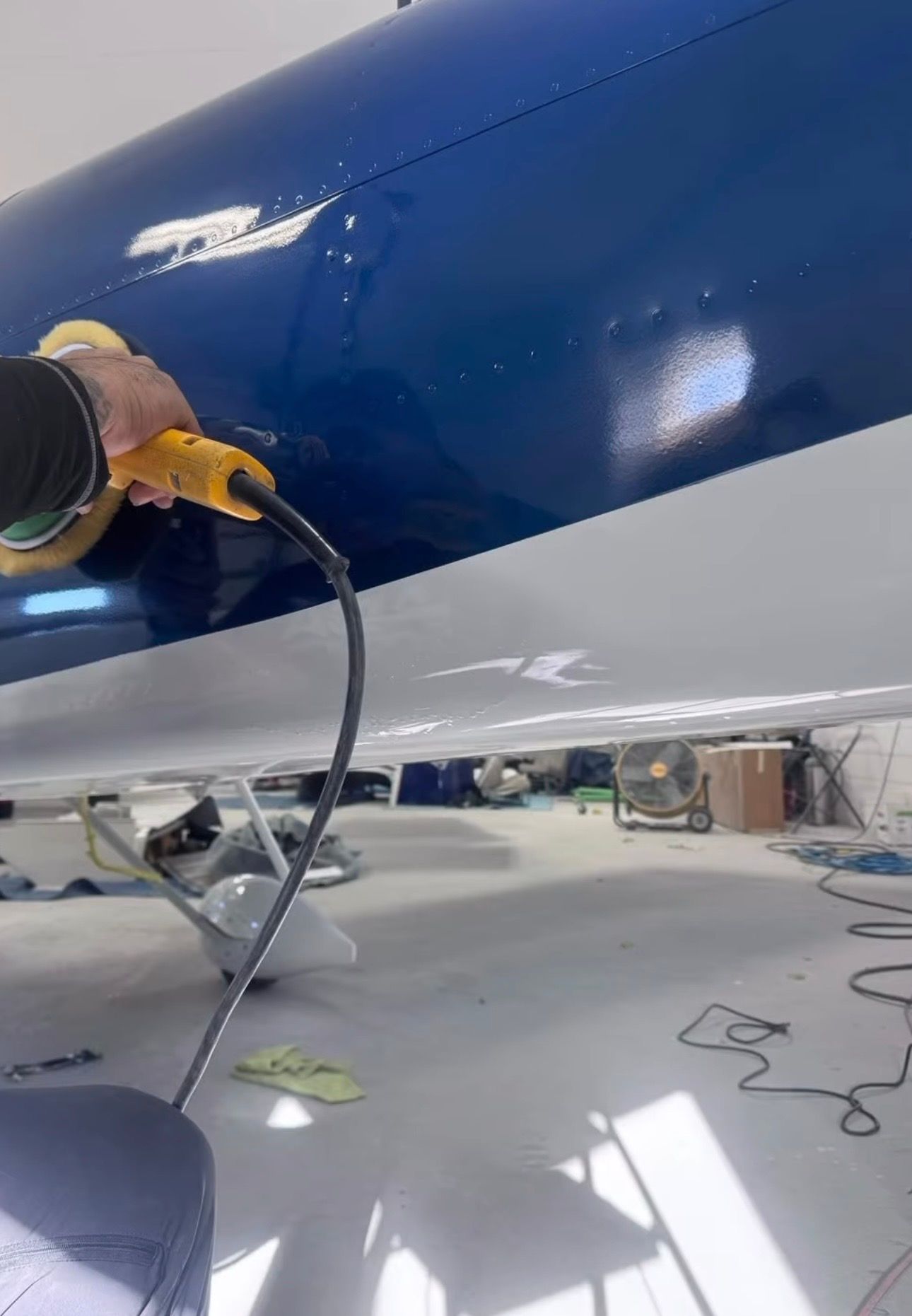 Hand polishing a blue and white boat hull in a workshop, with a power buffer in use