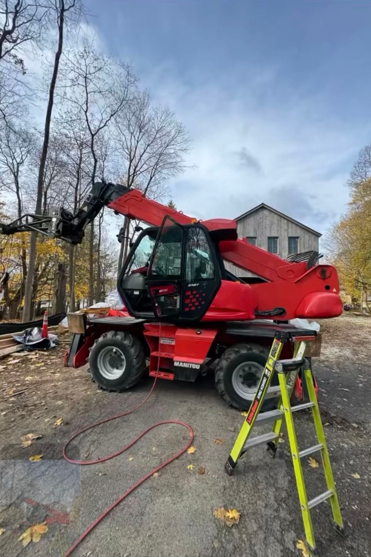 Red telescopic handler with raised boom in a wooded yard, with a ladder in front and a house behind.