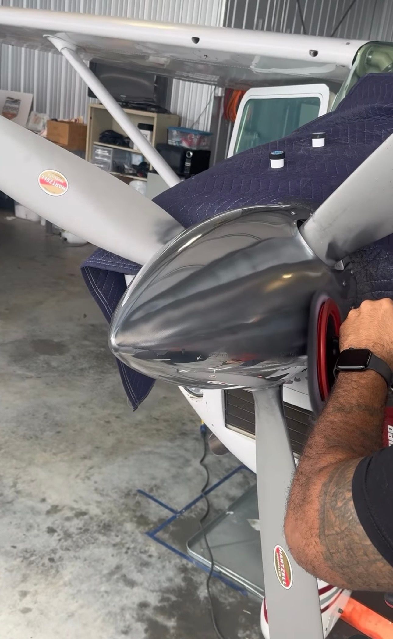 Close-up of a small plane’s nose and propeller in a hangar, with a person’s arm at right.