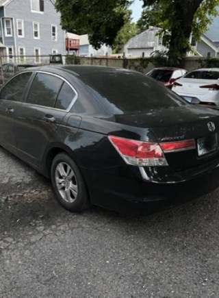 Black sedan parked on a gravel lot beside residential buildings and trees.