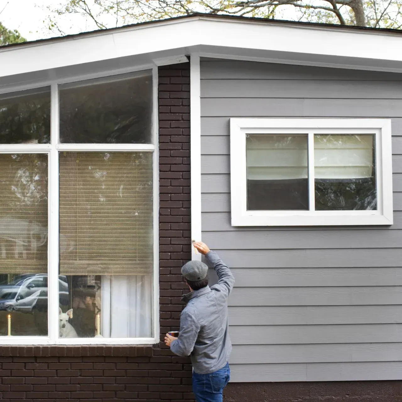Person painting the exterior of a gray house near a large window and brick trim