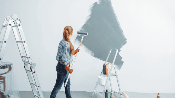 Person painting a large gray wall in a bright room with a ladder and paint supplies nearby