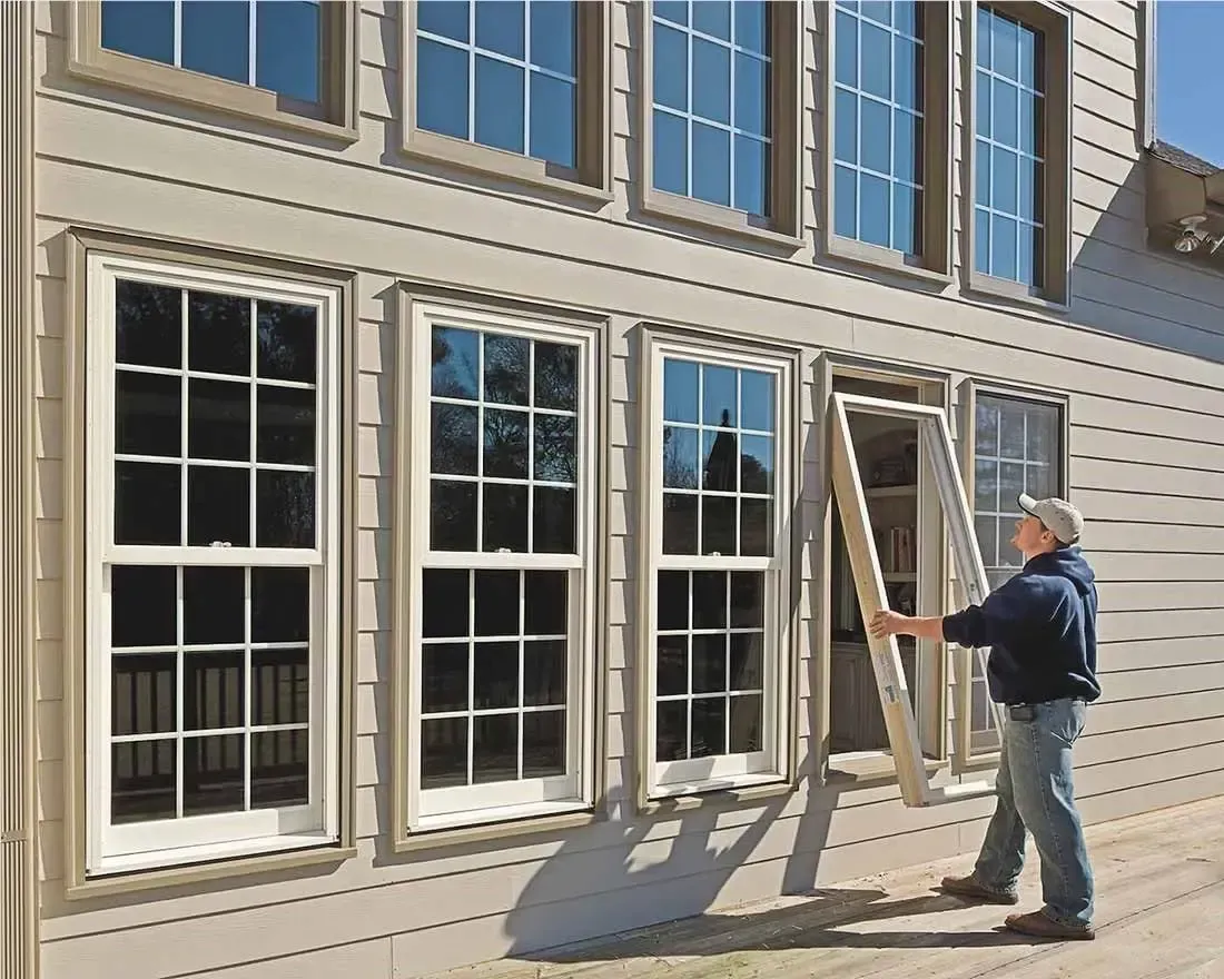 Worker carrying window frame beside beige house with tall windows and siding