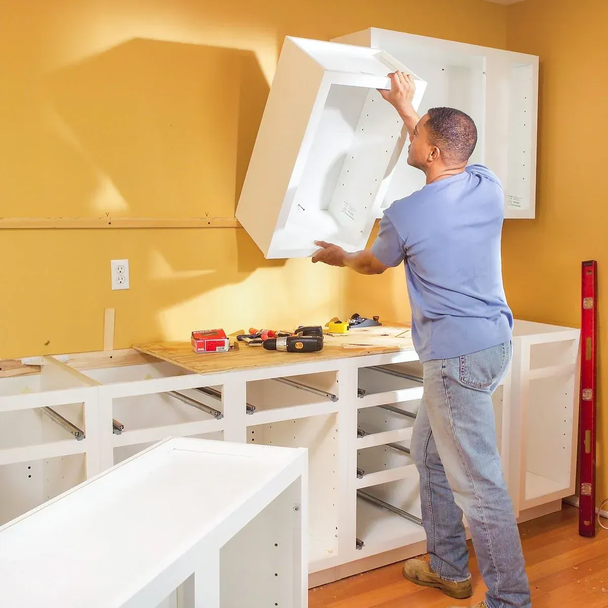 Person installing a white cabinet in a bright kitchen with yellow walls and open base cabinets