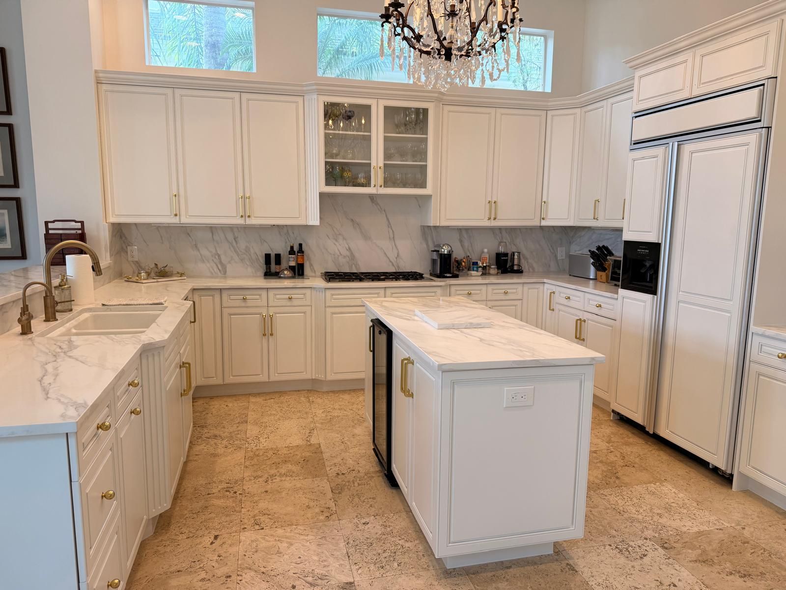 Bright white kitchen with marble island, cabinets, chandelier, and stainless steel fridge and sink.