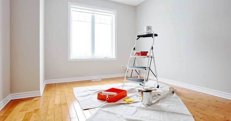 Empty room with hardwood floor, window, and paint supplies on a drop cloth, ready for renovation