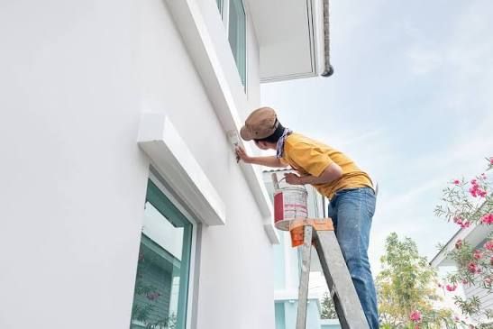 Person on a ladder painting a white exterior wall with a roller
