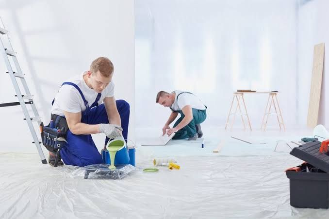 Two painters in white overalls working in a bright room with paint buckets and protective floor covering