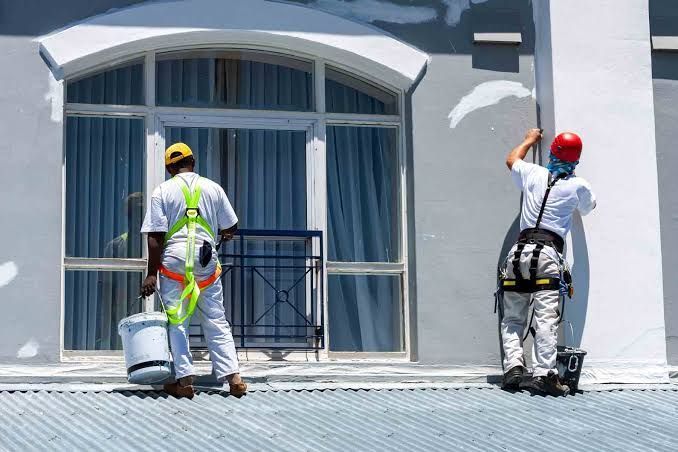 Two construction workers painting a white building exterior, one on a ladder and one on the ground