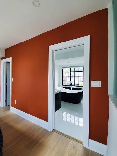 White hallway with orange wall, hardwood floors, and an open doorway into a bright room with a black chair.