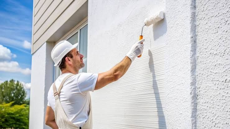 Painter in white overalls painting a white exterior wall with a roller on a sunny day