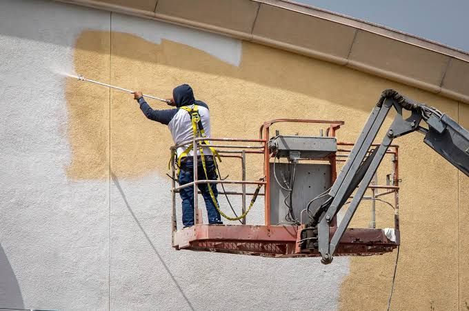 Worker spraying tan paint from a lift onto a building wall beside unfinished white stucco