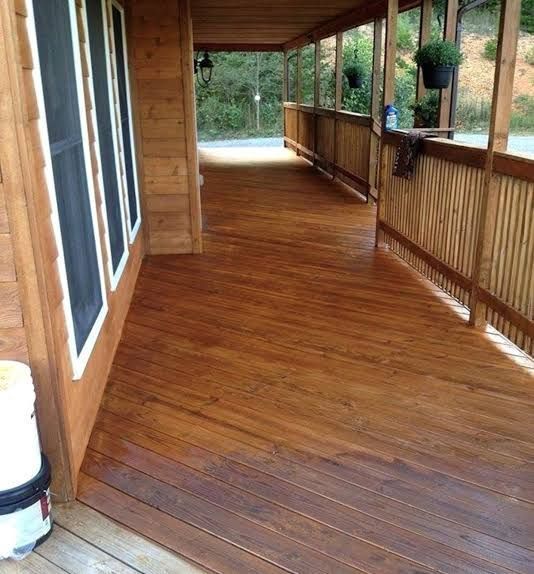 Covered wooden porch with railing, dark-stained floor, and windows along the left side.