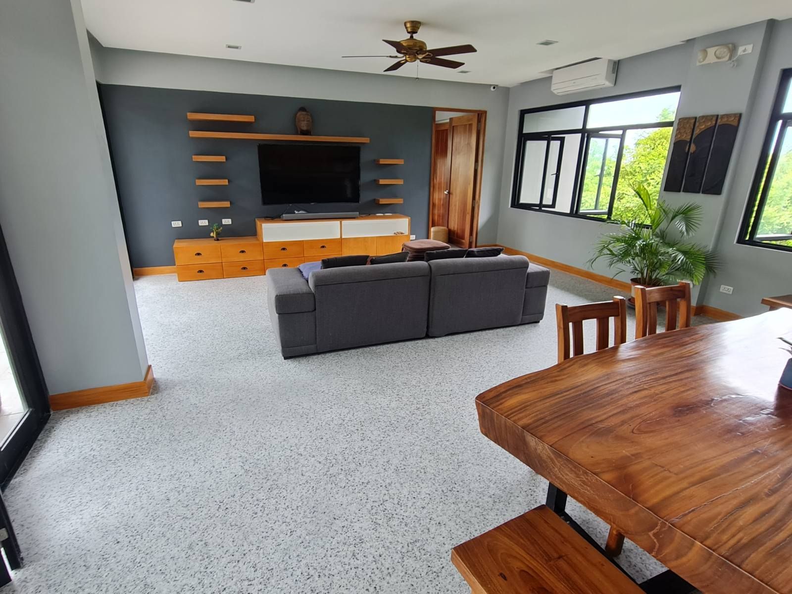 Modern living room with gray sofa, wooden TV wall unit, large windows, and dining table in foreground