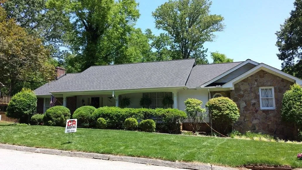 Single-story house with stone facade and dark gray roof, lush green lawn, and shrubbery in front.