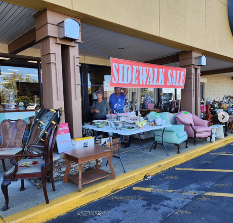 Sidewalk sale outside a store with tables of goods and a red banner, under a covered walkway. Sidewalk sale outside a store with tables of goods and a red banner, under a covered walkway.
