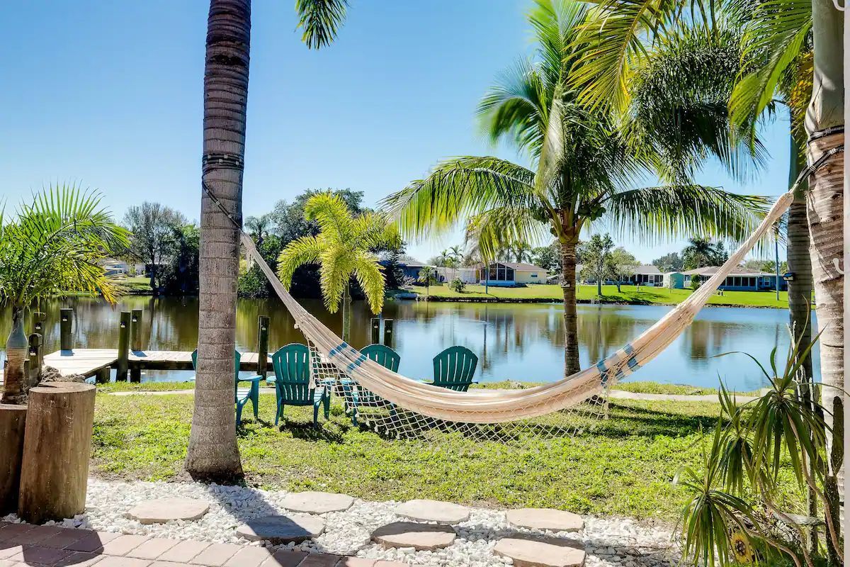 Palm trees and a hammock beside a sunny lakeside yard with grass and a patio.