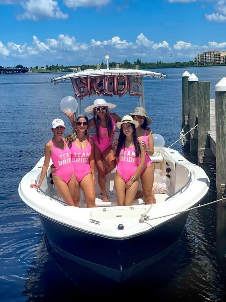 Five women in pink swimsuits on a white boat in blue water, holding a “BRIDE” sign.