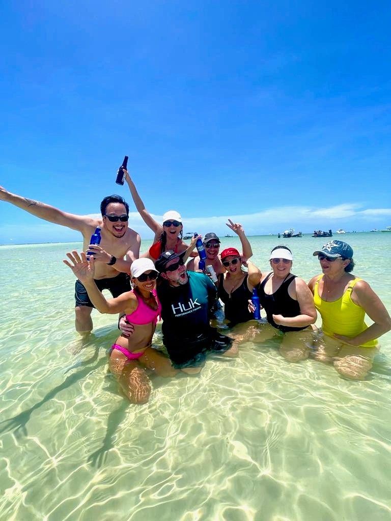 Group of friends posing and smiling in clear shallow ocean water under a bright blue sky.